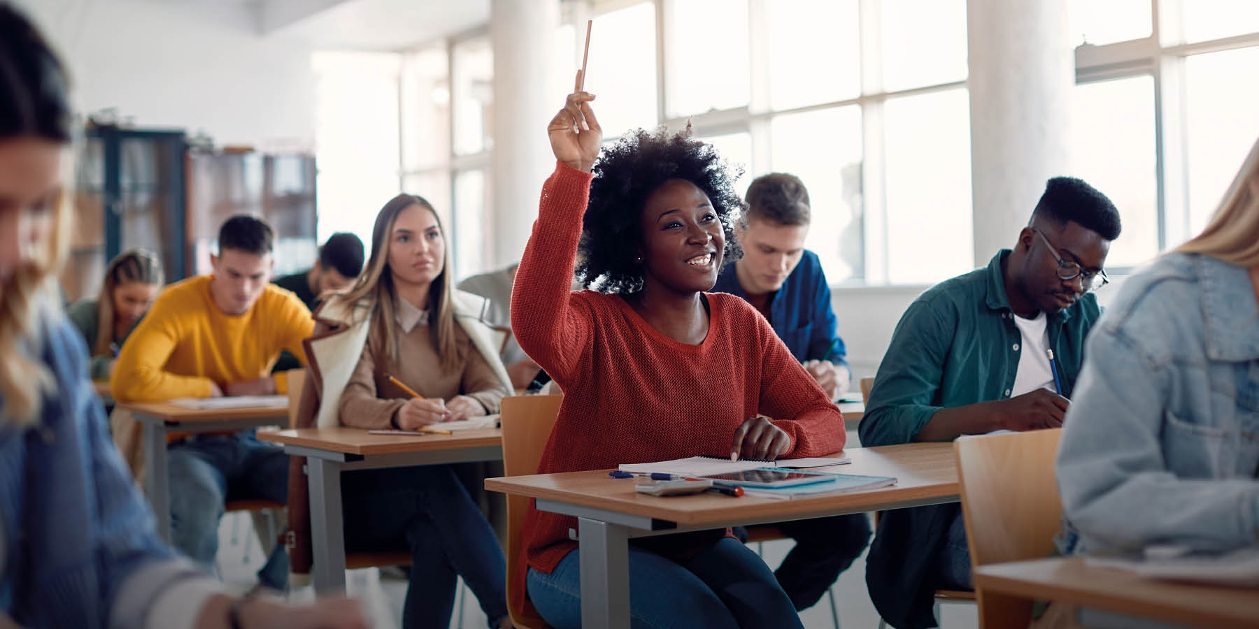 Klassenzimmer mit Schülern, die an Schreibtischen sitzen. Eine Person im Vordergrund hebt die Hand.
