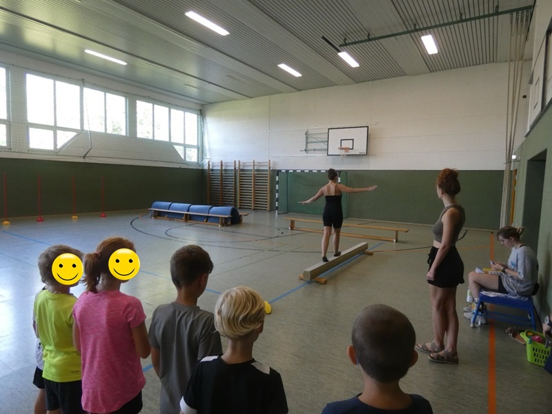 Kinder stehen in der Sporthalle und schauen einer Auszubildenden zu, die auf einem Schwebebalken balanciert. 