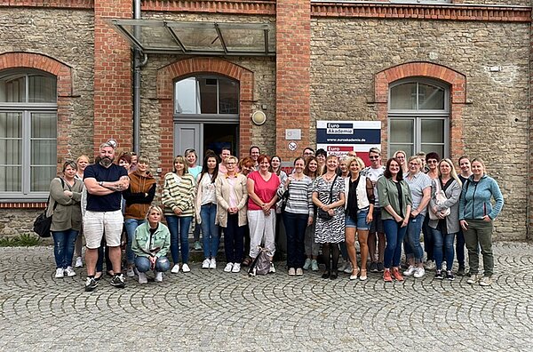 Eine Gruppe von Personen steht vor einem Backsteingebäude mit großen Fenstern und einem bogenförmigen Eingang. Ein Schild neben der Tür trägt die Aufschrift “Euro Akademie".”.