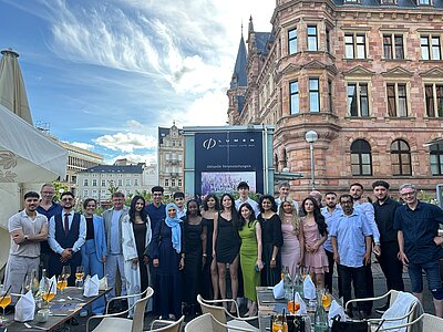 Eine große Gruppe festlich gekleideter junger Menschen und einige Erwachsene posieren gemeinsam im Außenbereich eines Restaurants vor einem historischen Gebäude. Die Stimmung ist feierlich, die Tische sind mit Getränken und Gedecken vorbereitet.