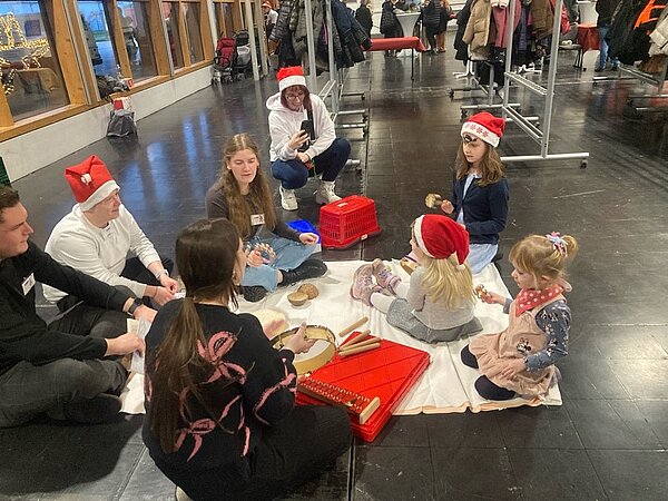 Weihnachtszeit fröhliche Zeit? Eine Gruppe von Erwachsenen und Kindern sitzt im Kreis auf dem Boden und spielt mit verschiedenen Musikinstrumenten. Alle tragen Weihnachtsmützen, und im Hintergrund sind Kleiderständer zu sehen.