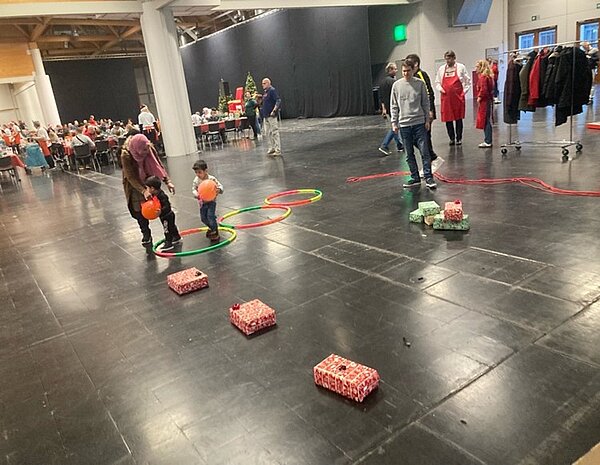 Weihnachtszeit fröhliche Zeit? Einige Kinder und Erwachsene nehmen an einer Weihnachtsveranstaltung teil, bei der in einer großen Halle ein Parcours mit Geschenkpäckchen und bunten Kreisen aufgebaut ist. Eine Erwachsene begleitet ein kleines Kind, welches einen orangefarbigen Luftballon hält. Im Hintergrund sind lange Tischreihen, Stühle zu sehen, auf denen Menschen sitzen. Daneben steht ein Weihnachtsbaum.