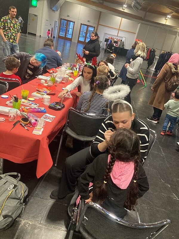 Weihnachtszeit fröhliche Zeit? Eine Gruppe von Kindern und Erwachsenen sitzen an einem langen Tisch. Die Erwachsenen schminken die Kinder. Im Hintergrund sind weitere Erwachsene und weitere Kinder zu sehen, die sich in einem Gemeinschaftsraum aufhalten. Der Tisch ist mit Schminkutensilien bedeckt.