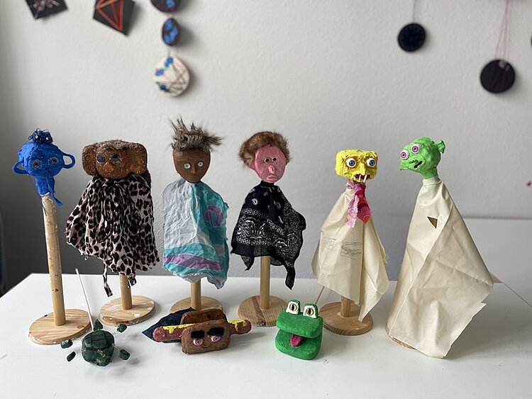 Projekt Schule ohne Rassismus Auf einem Tisch stehen verschiedene Handpuppen auf Holzständern. Diese haben grüne, gelbe, rote, schwarze und blaue Köpfe und jeweils Körper aus Stofffetzen, darunter einen mit Leopardenmuster.