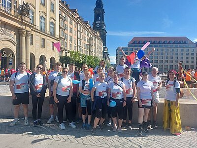 Eine Gruppe von Männern, Frauen und Kindern in Laufoutfits steht auf einem Platz, im Hintergrund ist ein Kirchturm zu sehen. Einige Personen halten bunte Luftballons in der Hand. Blauer Himmel, die Sonne scheint.