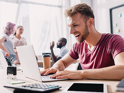 Ein lächelnder Mann mit Bart und rotem T-Shirt sitzt an einem Schreibtisch und arbeitet an einem Laptop. Auf dem Tisch befinden sich ein Taschenrechner, ein Stempel und ein Kaffeebecher. Im Hintergrund sind mehrere Personen in einem Büroumfeld zu sehen, die sich unterhalten.