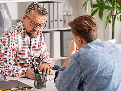 Zwei Männer sitzen sich an einem Schreibtisch in einer Büroumgebung gegenüber, während der ältere der beiden Männer auf ein Papier schreibt. Der jüngere Mann ist nur von hinten zu sehen.