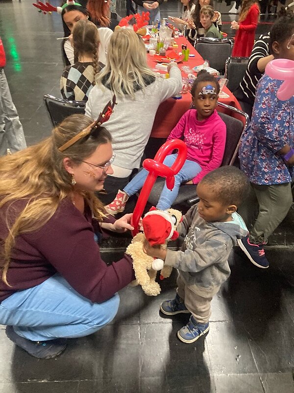 Weihnachtszeit fröhliche Zeit? Eine Frau kniet neben einem kleinen Jungen und gibt ihm ein Stofftier mit einem roten Hut und einem roten Ballon in Form eines Schlüssels. Im Hintergrund sitzen mehrere Kinder an einem Tisch und scheinen an einer Veranstaltung teilzunehmen.