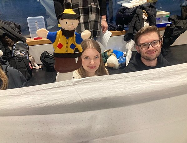 Weihnachtszeit fröhliche Zeit? Zwei Personen, eine Frau und ein Mann, hocken hinter einem weißen Vorhang und schauen lächelnd darüber. Die Frau hält eine Kasperle-Handpuppe hoch. Im Hintergrund liegen Jacken und Taschen herum.