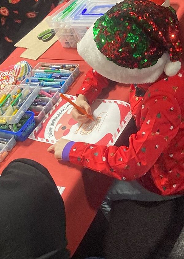 Weihnachtszeit fröhliche Zeit? Ein Kind in einem roten Weihnachtspullover und einer Weihnachtsmütze sitzt an einem Tisch und malt ein Bild mit Buntstiften. Auf dem Tisch liegen verschiedene Malutensilien.
