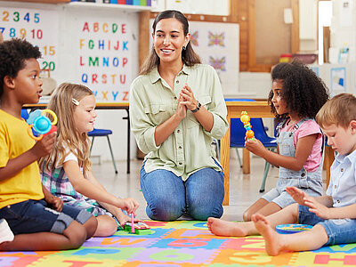 Auf dem bunten Teppich im Klassenzimmer sitzen Kinder und ein Erwachsener. Die Wände sind mit Buchstaben und Zahlen dekoriert.