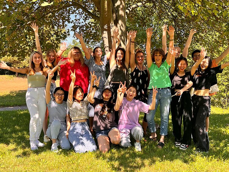 Eine Gruppe von 14 fröhlichen Menschen, hauptsächlich Frauen, posiert unter einem Baum auf einer grünen Wiese. Sie heben die Arme in die Luft und lachen. Einige zeigen das Peace-Zeichen, andere strecken ihre Hände in die Höhe. Die Gruppe ist bunt gekleidet, mit verschiedenen Outfits von leger bis sommerlich schick. Die Sonne scheint, und der Hintergrund zeigt Natur mit Bäumen und Wiese.