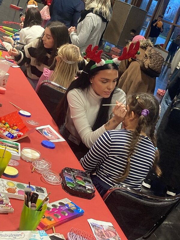 Weihnachtszeit fröhliche Zeit? Eine Frau malt einem kleinen Mädchen das Gesicht an. Sie sitzen an einem Tisch mit verschiedenen Schminkutensilien und Bastelmaterialien. Andere Kinder sind im Hintergrund zu sehen, und die Szene scheint Teil einer Veranstaltung oder eines Festes zu sein.