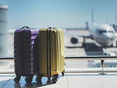 Zwei Rollkoffer, einer lila und einer gelb, stehen nebeneinander auf einem Flughafen mit einem Flugzeug im Hintergrund, was auf Reisen hinweist.